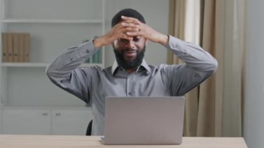 Odaklanmış genç sakallı zenci ofis çalışanı dizüstü bilgisayarla yazı yazıyor, ellerini başının arkasına koyuyor ve dikkati bilgisayardan dağılıyor. Modern odada temiz hava soluyun. 
