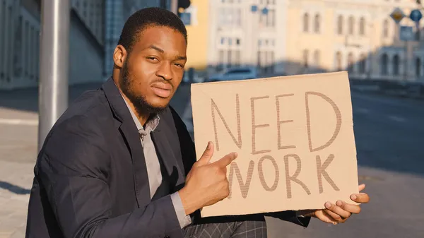 Triste raza mixta joven adulto negro hombre sentado al aire libre con tabla de texto de cartón Need Work. Busco trabajo. Despedido millennial molesto macho. Empresario que pierde negocio y busca empleo en crisis — Foto de Stock