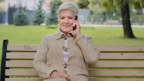 Usmívající se starší šedovlasá dáma s krásnou tváří sedí na lavičce v parku bere telefon, žena v důchodovém věku drží smartphone v ruce odpovědi na mobil má příjemný rozhovor s příbuznými — Stock fotografie