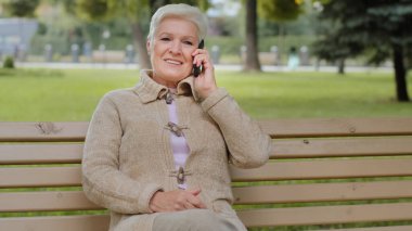 Parktaki bankta oturan mutlu, yüzü güzel yaşlı bir kadın telefon alıyor. Emeklilik yaşında, gri saçlı bir kadın cep telefonuyla cevap veriyor. Akıllı telefonu elinde tutuyor.