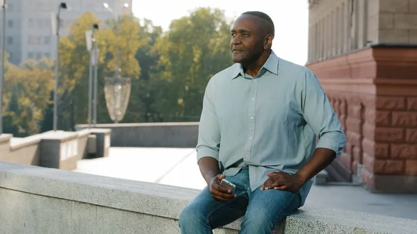 Stressad frustrerad äldre afroamerikansk man i pensionsålder i casual kläder skjorta och jeans sitter utomhus håller telefon väntar på klient eller vän sent för möte, ängsligt titta åt sidan — Stockfoto