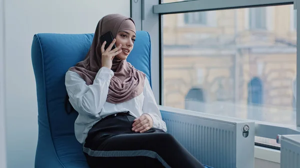 Sorridente Rilassato arabo manager millenaria donna in hijab seduto in poltrona confortevole parlare al telefono, consulenza, fare chiamate d'affari al lavoro, giovane lavoratrice o cliente che parla con il cellulare — Foto Stock