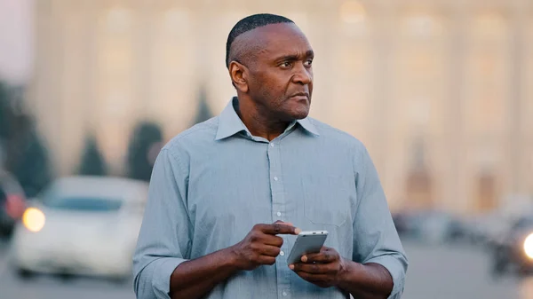 成熟的黑人男子拿着智能手机看着手机屏幕，在城市里寻找有电子地图的地址。老年商人非裔美国男性专业人员办公室大楼室外 — 图库照片