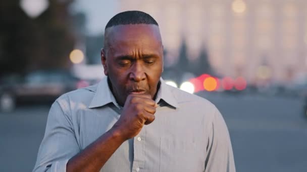 Зрелый африканский американец пенсионного возраста, стоящий на улице, страдающий бронхитом от кашля, кашляет астмой, держа в руках симптомы коронавируса респираторного вируса — стоковое видео