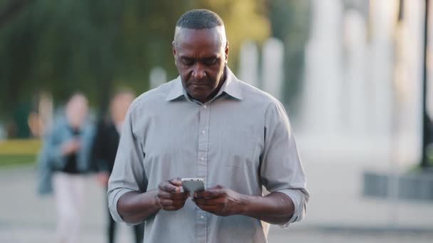 Verwirrter erwachsener afrikanischer Mann im Freien im Stadtpark, der sein Telefon in der Hand hält und sich verloren umsieht. Ältere schwarze Geschäftsmann versuchen, Gebäude mit Hilfe elektronischer Karte der Geolocation App im Smartphone zu finden — Stockvideo