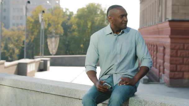Gefrustreerde Afro-Amerikaanse man met pensioen leeftijd in casual kleding shirt en jeans zitten buiten te wachten op klant of vriend laat voor de vergadering, angstig kijken naar smartphone scherm — Stockvideo