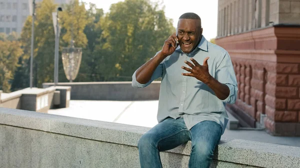 Worried nervous African American man in shirt and jeans holding mobile phone. Stressed frustrated client of retirement age talking to service sitting outdoors Customer dissatisfied with poor serve — Stock Photo, Image