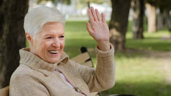 Lächelnde grauhaarige Oma in warmer Kleidung, die bei kühlem Wetter auf einer Bank sitzt, Hallo oder Auf Wiedersehen winkt, ältere Frau im Rentenalter, unbeschwertes Konzept — Stockfoto