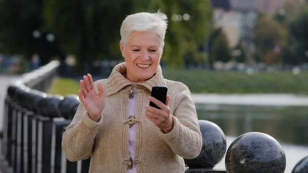 退職年齢の幸せなシニア女性は、スマートフォンのカメラを見て、ビデオ通話を行う。魅力的な古い成熟した女性は、携帯電話の屋外で話して良いニュースを伝える現代のガジェット無線インターネットを使用しています — ストック写真