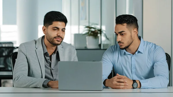 Dos hombres de negocios árabes jóvenes enfocados seriamente sentados en la oficina moderna usando el ordenador portátil que mira la pantalla de la computadora. Diversos empleados colegas de las Indias Orientales discuten trabajar juntos —  Fotos de Stock