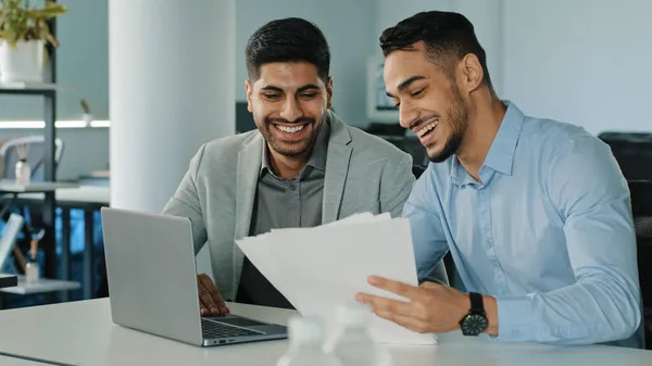 Jóvenes colegas sonrientes sosteniendo documentos en papel, empleados indios sentados en la oficina con computadora portátil recibiendo buenas noticias. Dos exitosos hombres de negocios árabes satisfechos excelente resultado de las negociaciones — Foto de Stock