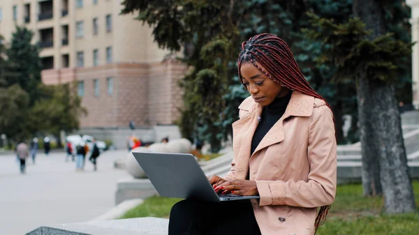 Poważny african amerykańska dziewczyna blogger za pomocą laptopa, siedzi na zewnątrz, młody zajęty skoncentrowany bizneswoman patrząc na ekranie, stażysta wpisując na klawiaturze, pisanie raportu, student pracuje nad projektem badawczym — Zdjęcie stockowe