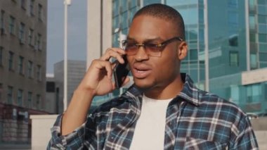 Moda gözlüklü mutlu Afrikalı adam şehirde cep telefonuyla konuşuyor. Yakın plan erkek, esmer tenli, konuşan akıllı telefon. Afro-Amerikalı adamın portresi. Uzaktan konuşma çağrısı.