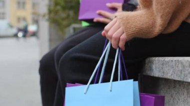 Bilinmeyen iki kişi ellerinde parlak alışveriş çantalarıyla sokakta oturuyorlar. Kara Cuma 'da indirimli indirimden sonra siyah pantolonlu kadın bacaklarını kapatıyorlar.