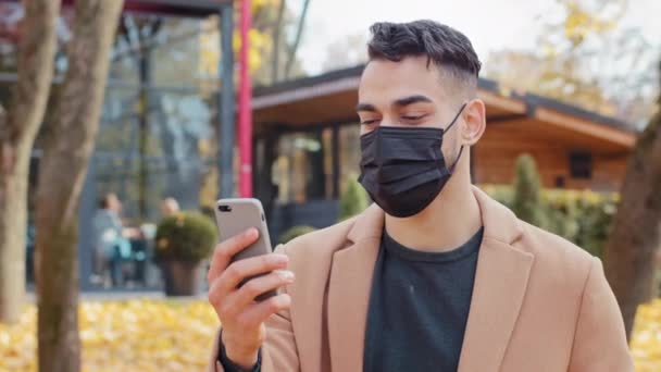 Hombre joven español en máscara médica hablando en videollamada usando teléfono inteligente de pie en el parque chico hispano asiente aprobadamente mirando la pantalla del teléfono al aire libre persona positiva agradable conversación en línea — Vídeo de stock