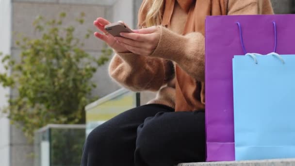 Jeune femme faisant du shopping en ligne le vendredi noir en utilisant le téléphone close-up fille méconnaissable consommateur assis dans la rue en ville avec des paquets lumineux achète messagerie féminine tenant téléphone mobile dans la main — Video