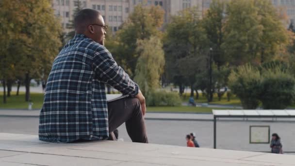 Zpět pohled africký Američan chlap obchodní muž vůdce v kostkované košile sedí na štěrku kroky město život na silnici mluví mobilní telefon vzdálená jednání volání odpovídání volání mobilní mluvení — Stock video