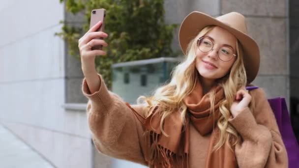 Felizmente mujer shopper respuesta video llamada chica atractiva hacer foto usando teléfono móvil mujer caucásica de pie en la calle con bolsas millennial rizado rubia dama con sombrero y gafas al aire libre — Vídeo de stock