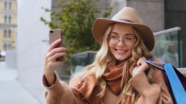 Приваблива дівчина-блогер знімає відео для блогу після покупки стоячи на вулиці в місті біла жінка фотографує на мобільній камері молоду тисячолітню леді, яка позує, тримаючи барвисті пакети на відкритому повітрі — стокове відео