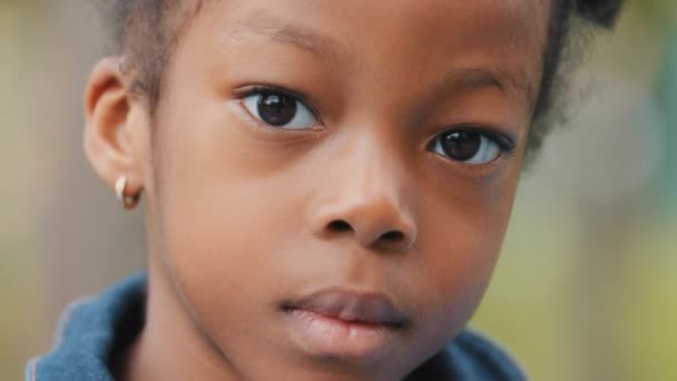 Close-up bez emocí dítě tvář africký americký holčička zamyšlený při pohledu na kameru smutný osamělý žádné emoce dítě vážné problémové dcera venku zadumaný pohled portrét roztomilé neusmívající se školačka — Stock video
