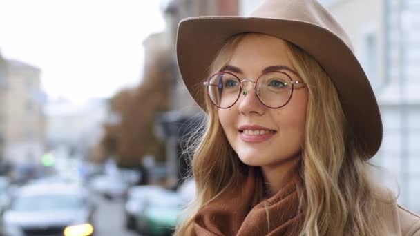 Fiatal gyönyörű kaukázusi lány áll a szabadban közelkép millenniumi nő kalap és szemüveg a város hátterében portré álmodozó nő mosolyog látszó távolság boldog szőke diák pózol az utcán — Stock videók
