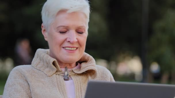 Heureuse femme âgée travaillant à l'ordinateur portable souriant femme âgée de l'âge de la retraite en utilisant l'ordinateur de navigation ou surfer sur Internet, lire des nouvelles en ligne, excité dame âgée textos message à PC assis à l'extérieur — Video