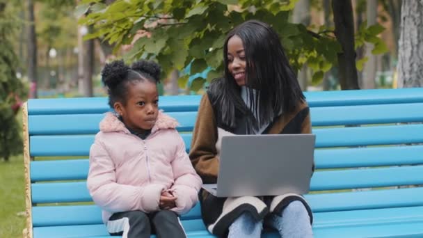 Genç Afro-Amerikan annesi, kızı ile birlikte şehir parkında bankta oturuyor. Diz üstü bilgisayar başında sevimli, siyah bir kız çocuğu, gülümseyen bir anneyle coşkulu bir şekilde sohbet ediyor. — Stok video