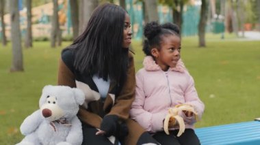 Genç anne ve kızın portresi, oyuncak ayıyla bankta oturmuş muz yiyor. Afro-Amerikan ailesinin açık havada eğlendiğini gösteriyor. Parkta gülümseyen sevimli bir çocukla.
