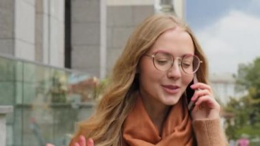 Şehirde yürüyen genç kız öğrenci telefonla konuşuyor. Yakın plan beyaz kadın arkadaşça cevaplar. Açık havada hareketli gülümseyen bayan sarışın, telefonda hoş sohbetlerden hoşlanıyor.