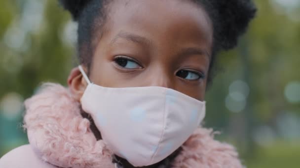 Gros plan mignonne petite fille masquée à l'extérieur enfant bouclé posant dans le parc de la ville portrait verrouillé grave enfant afro-américain seul debout dans la rue regardant la caméra épidémie covide pandémie coronavirus — Video