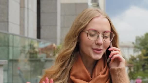 Ung flicka student promenader på gatan i staden tala på telefon närbild ansikte kaukasiska kvinna vänliga svar samtal leende aktivt gestikulera utomhus dam blond njuter av trevlig konversation på telefon — Stockvideo