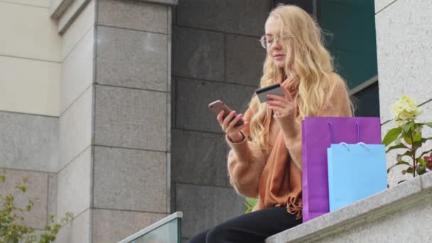 Attrayant acheteur féminin fait commande en ligne en utilisant téléphone femme caucasienne tenant la carte de crédit et entre le numéro pour la transaction dans l'application fille millénaire assis avec des sacs à provisions colorés près du bâtiment — Video