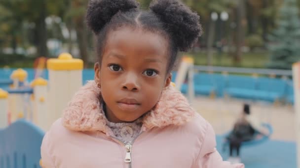 Allvarligt porträtt söt liten afro amerikansk unge inga känslor närbild ensam liten flicka tittar på kameran urban lekplats lugn känslomässigt barn efter skolan stående stadspark utomhus utan vänner — Stockvideo