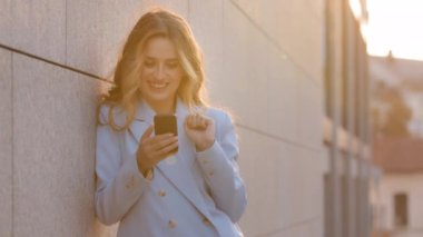 Güneşli arka planda duvarın yanında duran güzel, mutlu beyaz kız iş kadını cep telefonu sohbetlerine bakıyor akıllı telefonlar alıyor internetten mesaj yazma uygulaması kullanarak haberleri okuyor.