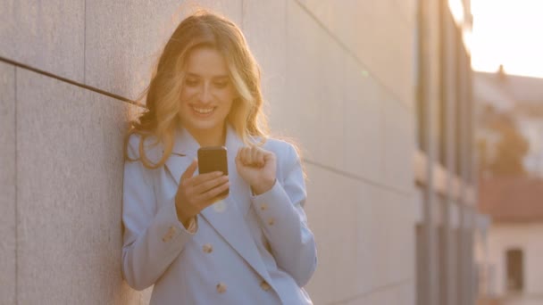 Belle caucasienne souriante heureuse femme d'affaires debout près du mur sur la rue en arrière-plan de soleil regardant dans le téléphone mobile chat smartphone acheter en ligne dactylographie message nouvelles en utilisant l'application — Video