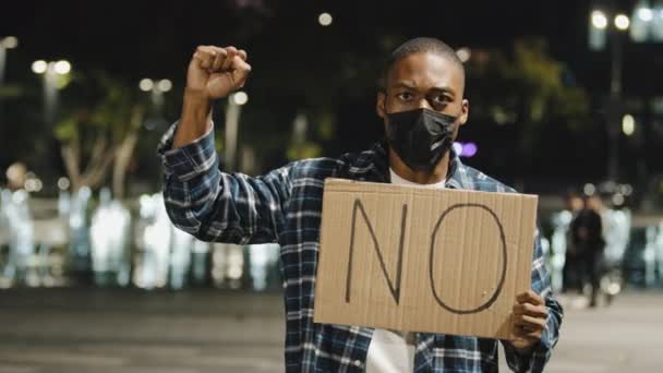 Africký Američan v lékařské masce stojí ve večerních hodinách město drží ceduli banner karton text ne, naštvaný chlap protestuje křičí mávání pěstí protestní aktivismus nesouhlas popírání černé životy záleží — Stock video