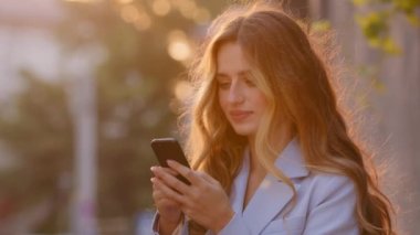 Gün batımında güneş ışığı altında sokakta dikilen güzel genç bir kadın cep telefonuna bakıyor internetten mesajları cevaplıyor akıllı telefonlardan sohbet ederek internetin keyfini çıkarıyor.