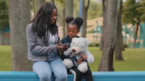 Позитивна мама і дочка сидять на лавці в парку мати з дитиною, посміхаючись дивлячись на телефон афроамериканська сім'я щасливо проводять час на відкритому повітрі маленька дівчинка тримає плюшевого ведмедя жінка використовує смартфон — стокове відео