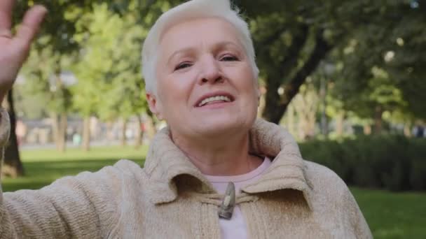 Retrato de una vieja jubilada de pelo gris feliz relajándose al aire libre saludando o despidiéndose. Abuela de mediana edad sonriente sincera disfrutando de una conversación de videollamada con amigos o niños mayores — Vídeo de stock