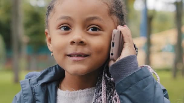 Маленькая девочка разговаривает по мобильному телефону на открытом воздухе портрет многорасовый ребенок смотрит в камеру отвечает на телефонный звонок серьезный африканский американец ребенок с косичками стоя на улице коммуникации использовать гаджет — стоковое видео