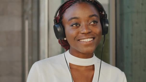 Portret van jonge mooie vrolijke grappige Afro-Amerikaanse meisje student toerist staat in koptelefoon luisteren naar muziek in de stad. Close-up vrouw zingen luistert naar audio geluiden zingen lied genieten — Stockvideo