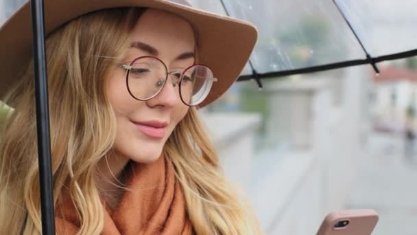 Close-up jong mooi vrouw onder paraplu glimlachen buiten student aantrekkelijk vrouw holding smartphone blank krullend blond meisje met hoed en bril chatten gebruik telefoon wachten op straat stad — Stockvideo