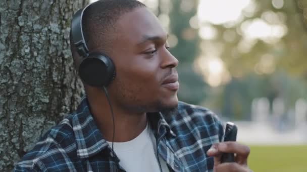 Hombre retrato afroamericano joven hombre divertido en auriculares escuchando música usando el teléfono en línea de radio pretendiendo tocar la batería canta en el teléfono inteligente como micrófono disfrutando de sonido de la canción de audio en el parque — Vídeos de Stock