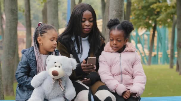 Afroamerikaner glücklich Familie sitzt auf Bank im Park Mutter mit Töchtern Blick auf Telefon kleines Mädchen hält Teddybär junge Frau kommuniziert mit Kindern Mutter und zwei Schwestern machen Videoanruf — Stockvideo