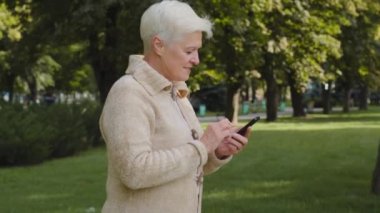Gri saçlı, emekli bir kadın, açık havada parkta yürüyor, telefon görüşmeleri yapıyor, akıllı telefon uygulamaları kullanıyor, ekrana bakıyor, son sınıf öğrencisi, internetten sohbet ediyor, alışveriş yapıyor, mobil cihazla haber okuyor.