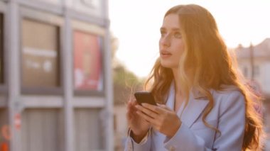 Güzel bir iş kadını sarışın kullanıcı gün batımında güneş ışığının altında durup mobil uygulama navigasyon kullanarak telefona bakıyor. Çevrimiçi haritada kendini işaret ederek konuşuyor.