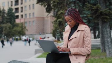 Ciddi bir Afro-Amerikan kız blogcusu dizüstü bilgisayar kullanıyor, dışarıda oturuyor, ekrana bakmakla meşgul genç bir iş kadını, klavyede staj yapıyor, rapor yazıyor, araştırma projesi üzerinde çalışıyor.