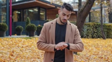 Sonbahar parkında sabırsız İspanyol yakışıklı bir adam buluşmayı bekliyor hoşnutsuzca kol saatine bakıyor ve dışarıda dikilirken kafasını sallıyor sinirli sakallı bir erkek.