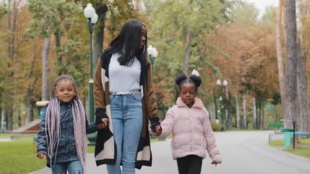 Jovem mãe segurar duas filhas por mãos família afro-americana feliz amigável andando no parque da cidade no fim de semana meninas pequenas e mãe desfrutar de andar ao ar livre mulher com crianças passar o tempo juntos sorrindo — Vídeo de Stock