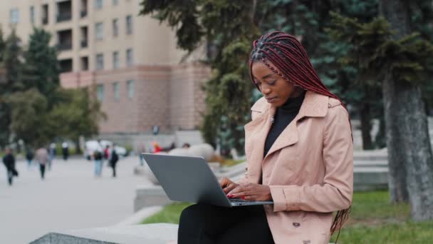 Ernste afrikanisch-amerikanische Bloggerin mit Laptop, im Freien sitzend, junge, beschäftigte Geschäftsfrau, die auf den Bildschirm schaut, Praktikantin, die auf der Tastatur tippt, Bericht schreibt, Studentin, die an Forschungsprojekt arbeitet — Stockvideo